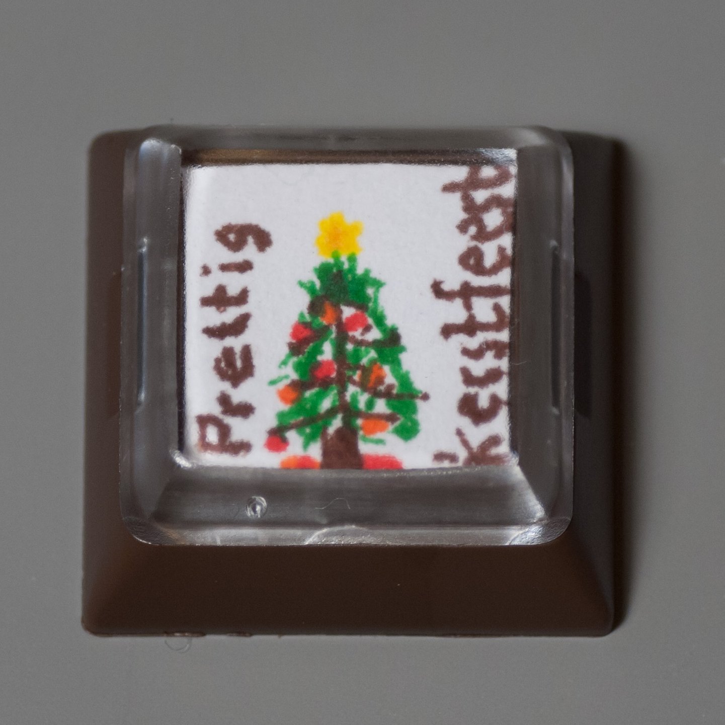 This is a relegendable DSA keycap. All craftsmanship is of the highest qualitiy. The base is shot in brown ABS, the top is molded from transparent polycarbonate. Embedded in between is a masterfully decorated piece of paper by Secret Santa.<br />On the item is an image of a Christmas tree in brown and green. The tree is decorated with red and orange balls. It is menacing with a yellow, star-formed spike. The item is inscribed with the words "prettig kerstfeest" in brown ink.<br />The artwork relates to the celebrating of Christmas in the Netherlands in late December of 2013.
